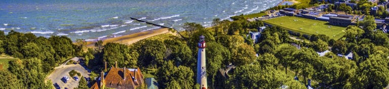 Aerial view of a coastal area with a lighthouse, beach, red-roofed building, and surrounding greenery.