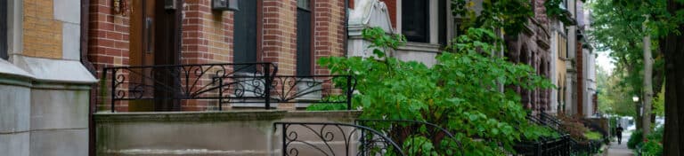 A row of brick townhouses with black wrought iron railings and green shrubs along the sidewalk.