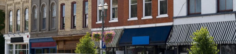 Row of small-town storefronts with varied architecture, awnings, trees, and a streetlamp with flower baskets.