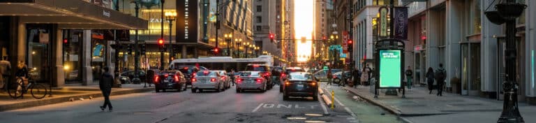 Busy urban street at dusk with cars, buses, pedestrians, and illuminated signs.