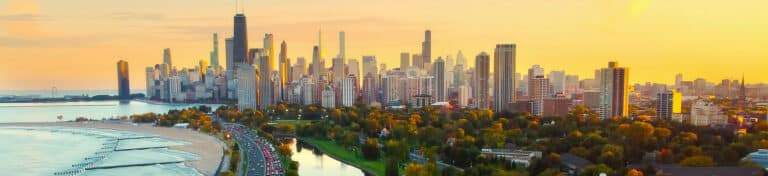 Panoramic view of Chicago skyline at sunset with Lake Michigan, beach, and tree-lined road.
