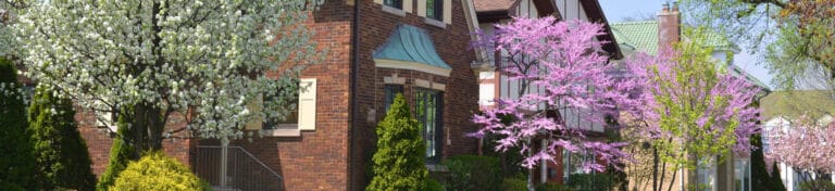 Row of brick houses with blooming trees and gardens in a springtime neighborhood.