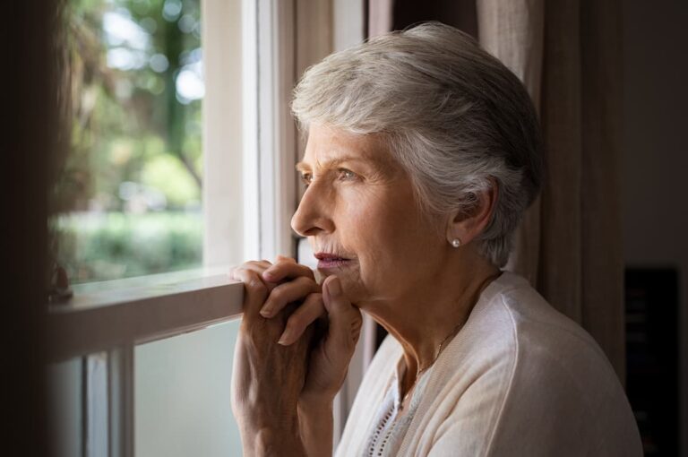 An older woman gazes thoughtfully out of a window, reflecting on the view outside.