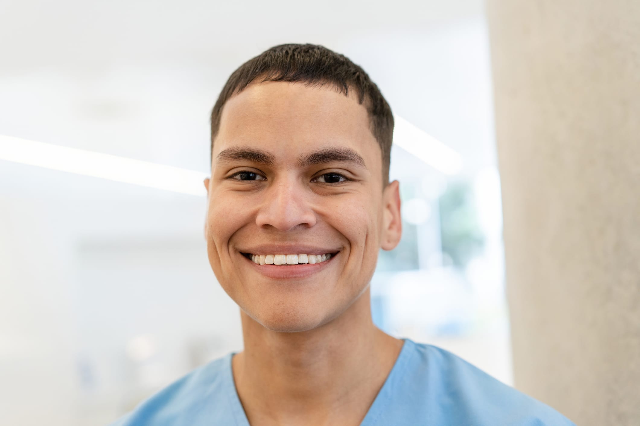 Close-up portrait of a happy young male nurse standing in hospital. Confident healthcare worker wearing blue scrubs looking at camera.